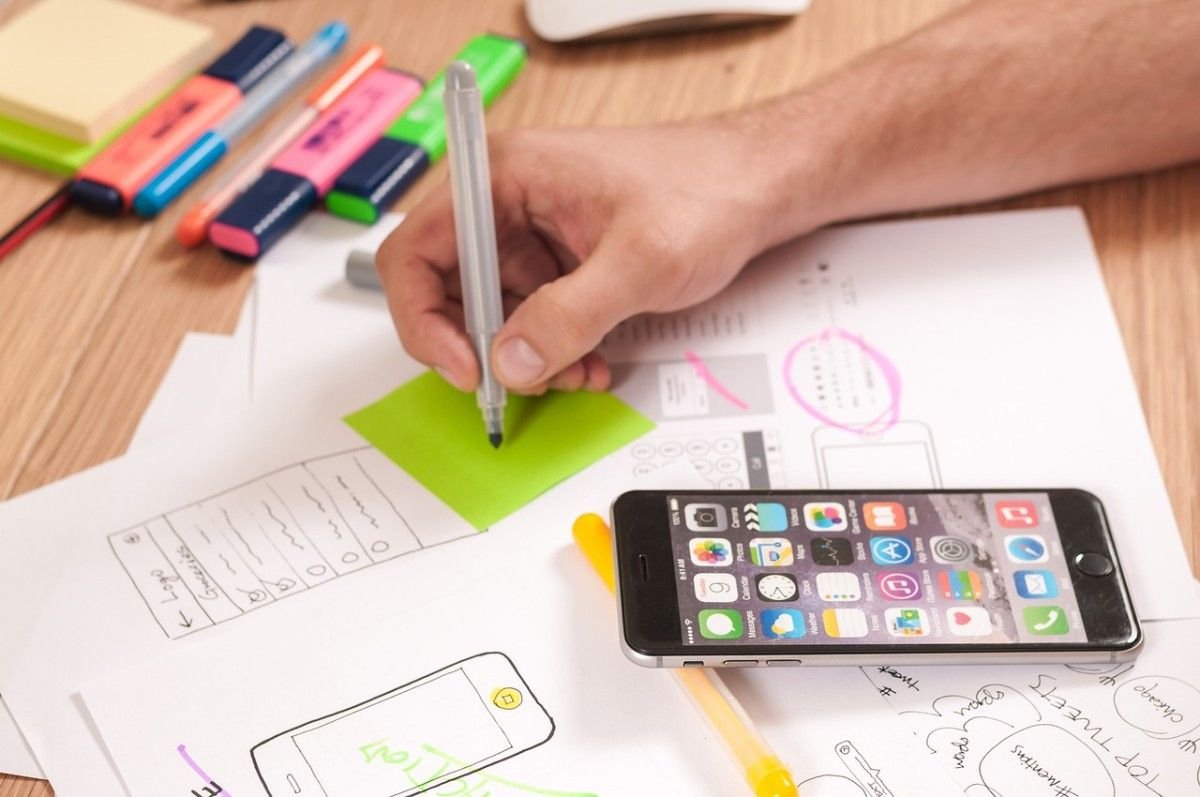 A person writes on a green sticky note at a cluttered desk with smartphone, UX/UI sketches, colored pens, highlighters, and paper clips. The phone rests on top of wireframe drawings for a mobile app.